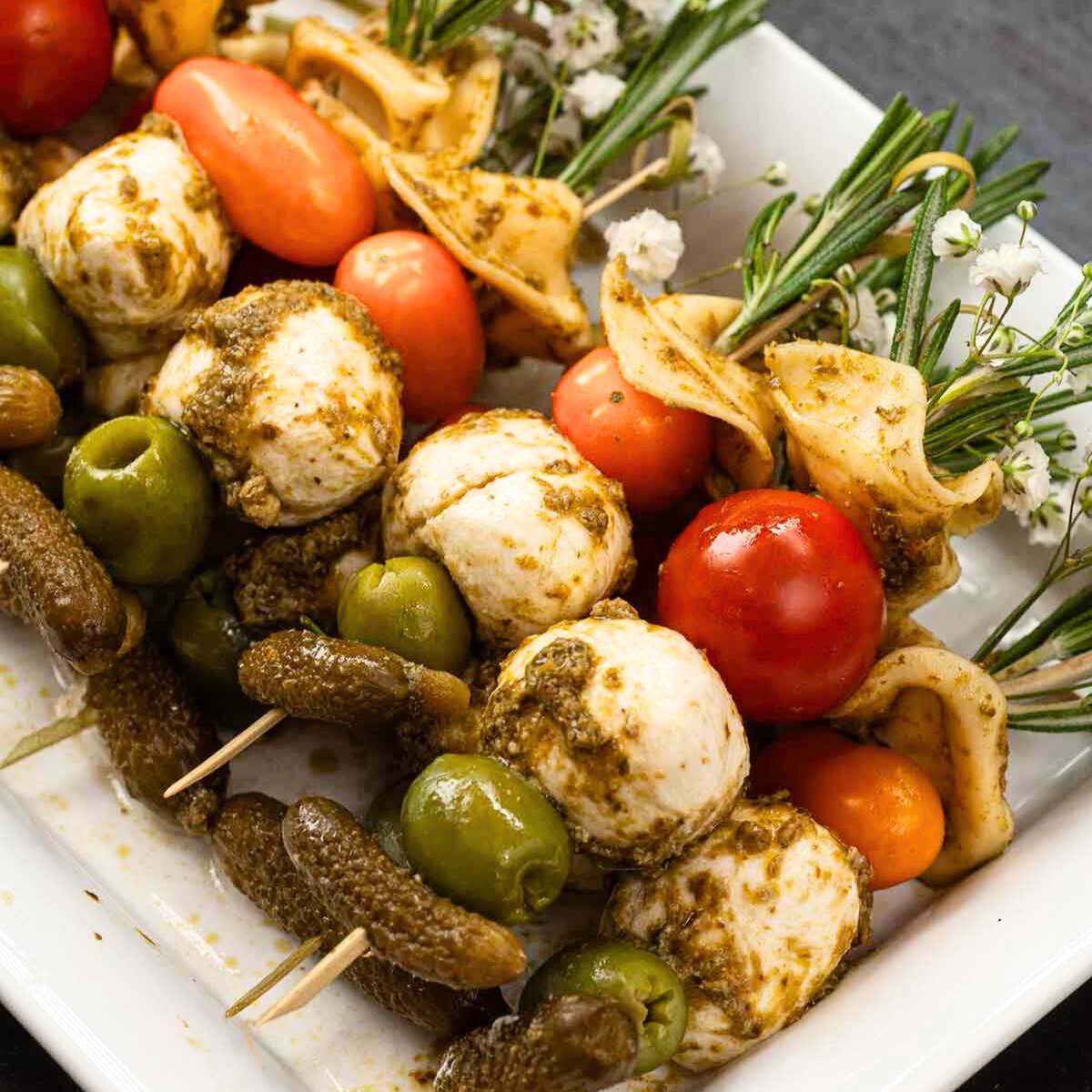 A platter of skewered appetizers with mozzarella balls, cherry tomatoes, olives, and pickled vegetables, drizzled with pesto sauce.