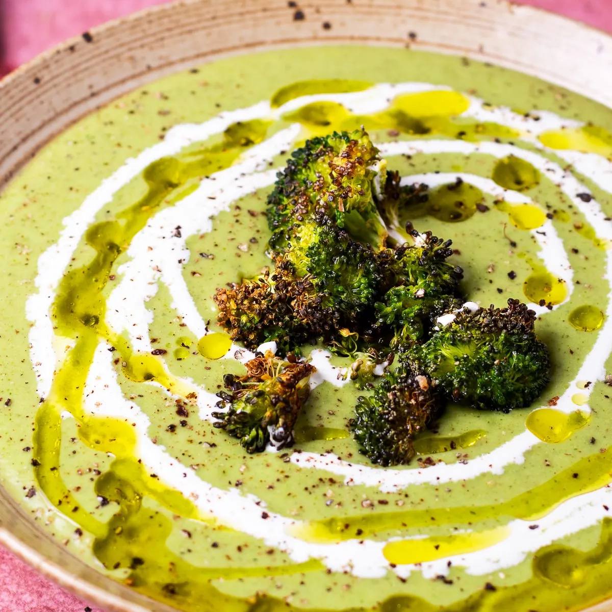 A bowl of creamy green broccoli soup topped with roasted broccoli florets. The soup is drizzled with olive oil and cream.