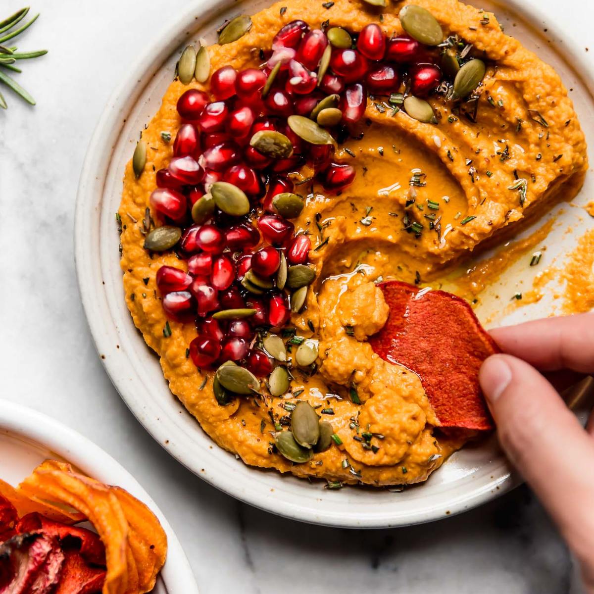 A bowl of creamy pumpkin hummus topped with pomegranate seeds, pumpkin seeds, and rosemary. A hand is dipping a sweet potato chip into the hummus.