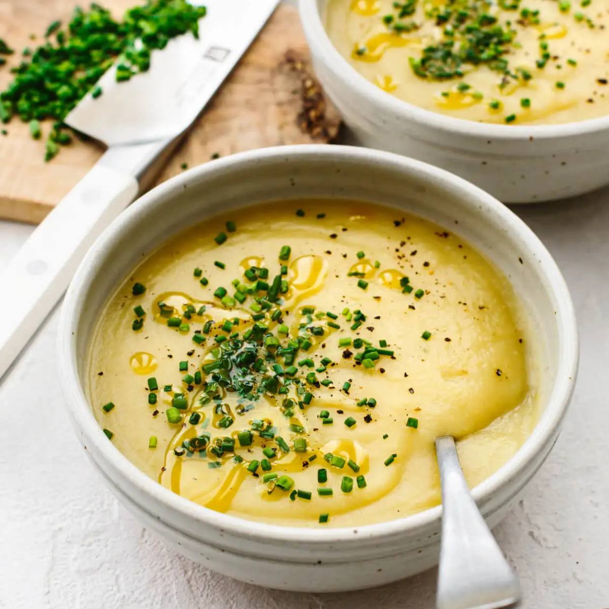 A white bowl filled with a creamy potato soup. The soup is topped with chives, black pepper, and a drizzle of olive oil. A spoon is resting in the bowl.