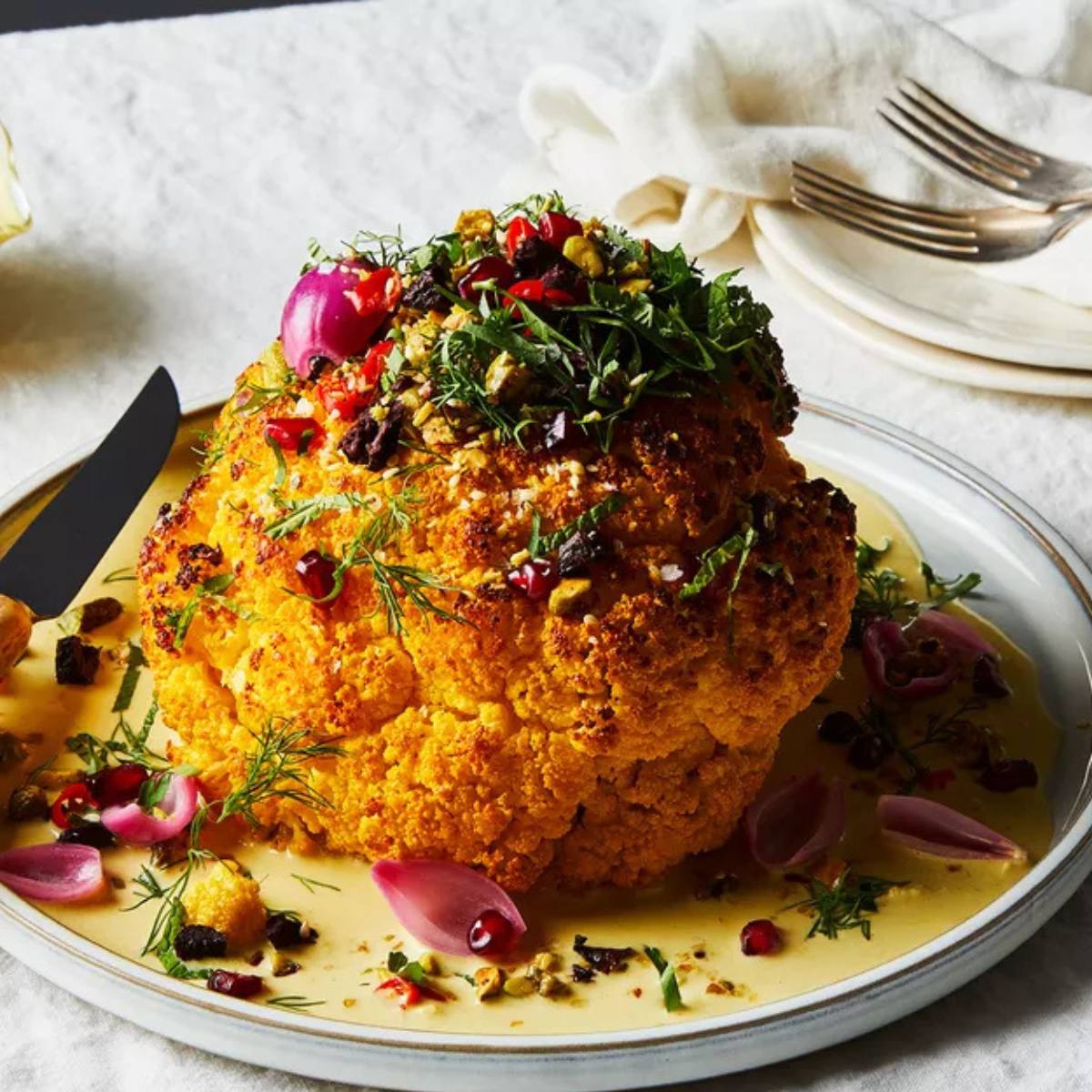 A golden-brown roasted whole cauliflower topped with a colorful mixture of herbs, pomegranate seeds, and pistachios.