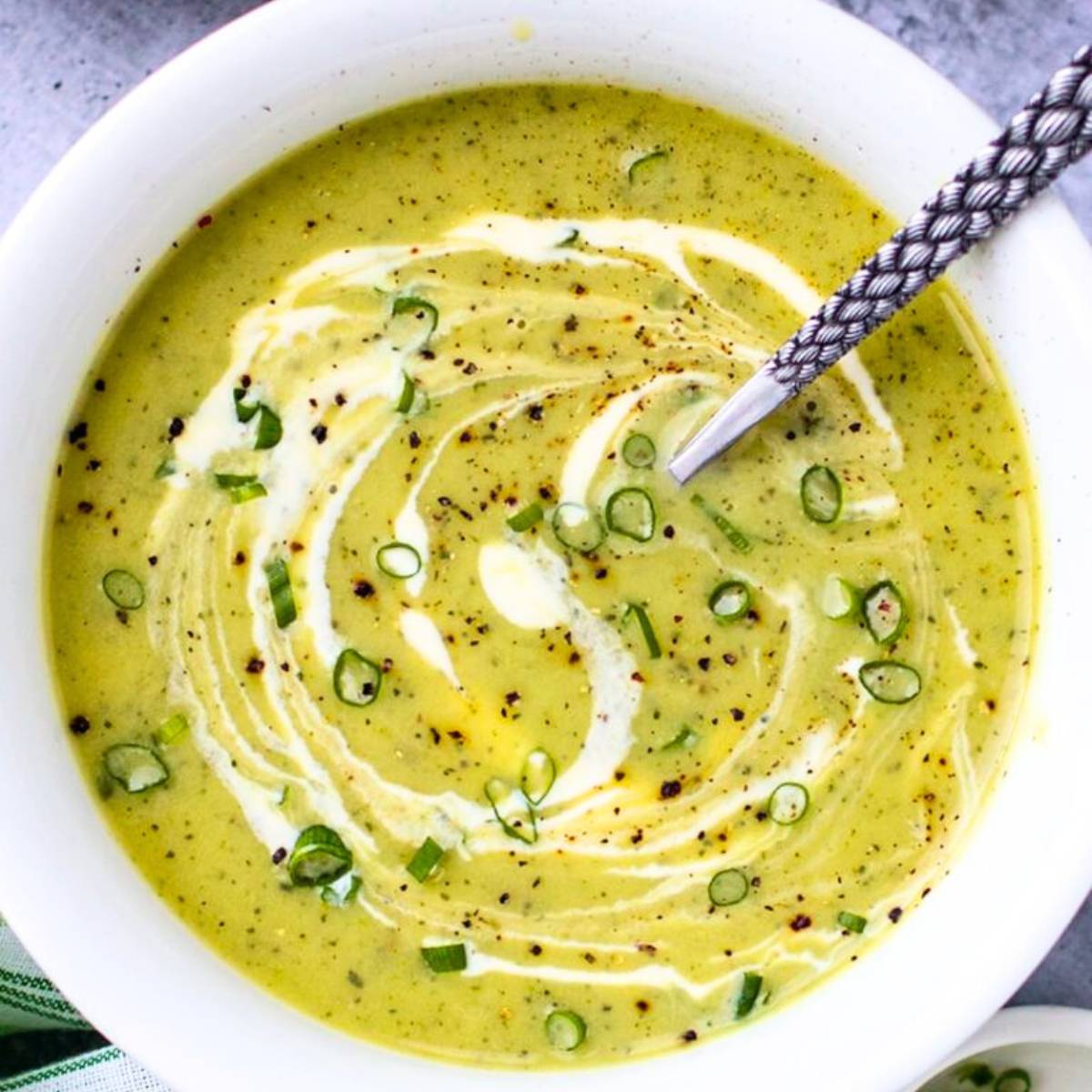 A bowl of creamy green soup topped with dollops of sour cream, chopped chives, and a sprinkle of black pepper.