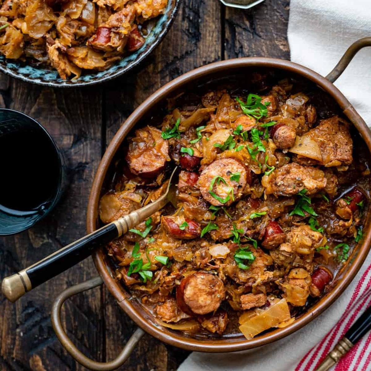 A copper pot filled with a savory stew of sauerkraut, sausage, and meat. The stew is garnished with fresh herbs. A plate of additional sauerkraut and a glass of wine are visible in the background.