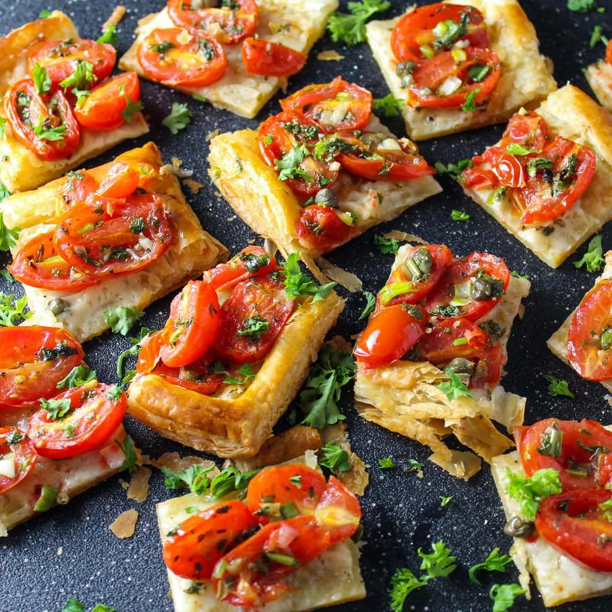 A close-up of several small, golden-brown puff pastry squares topped with roasted cherry tomatoes, capers, and fresh herbs.