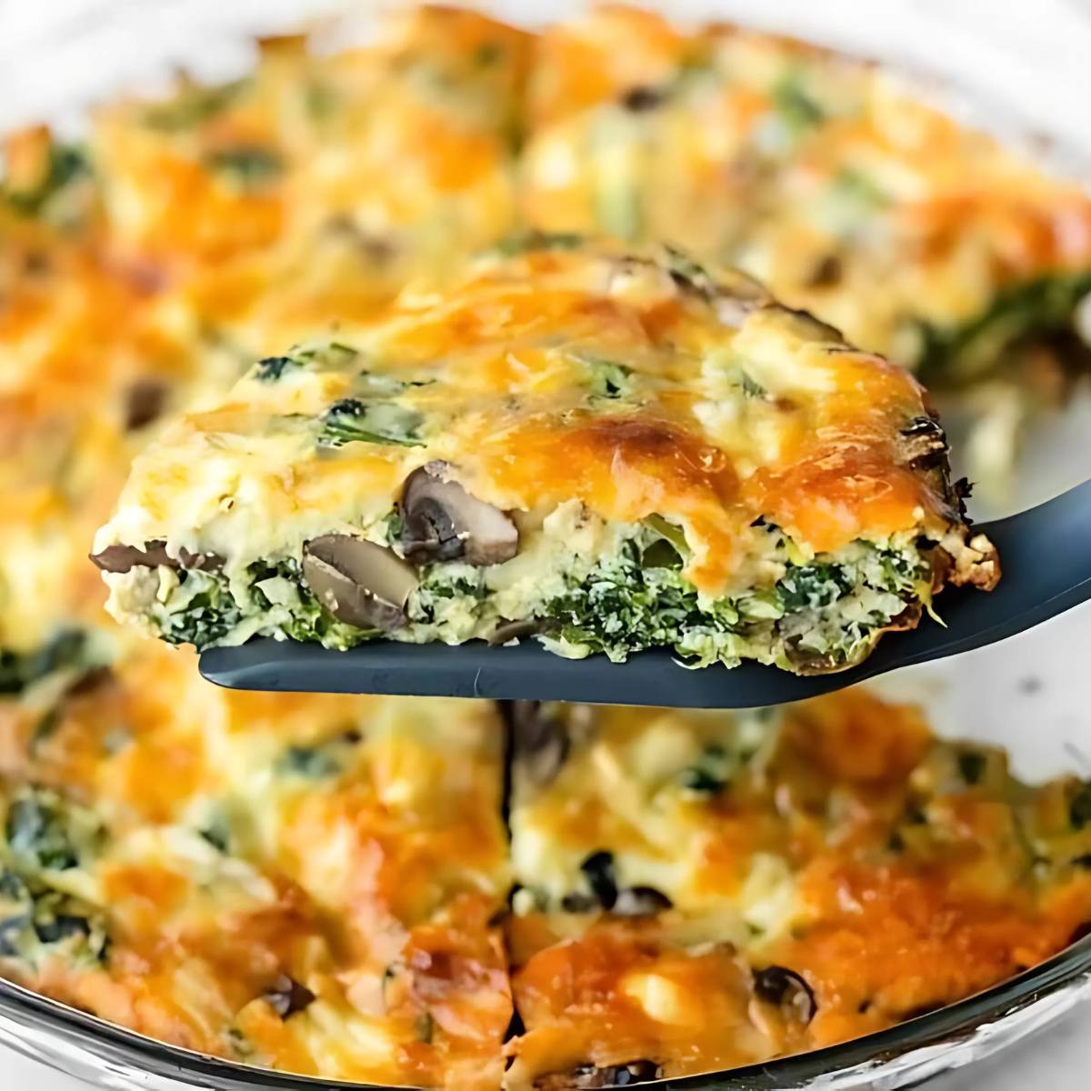 A golden-brown slice of quiche with spinach, mushrooms, and cheese being lifted from a pie dish with a spatula.