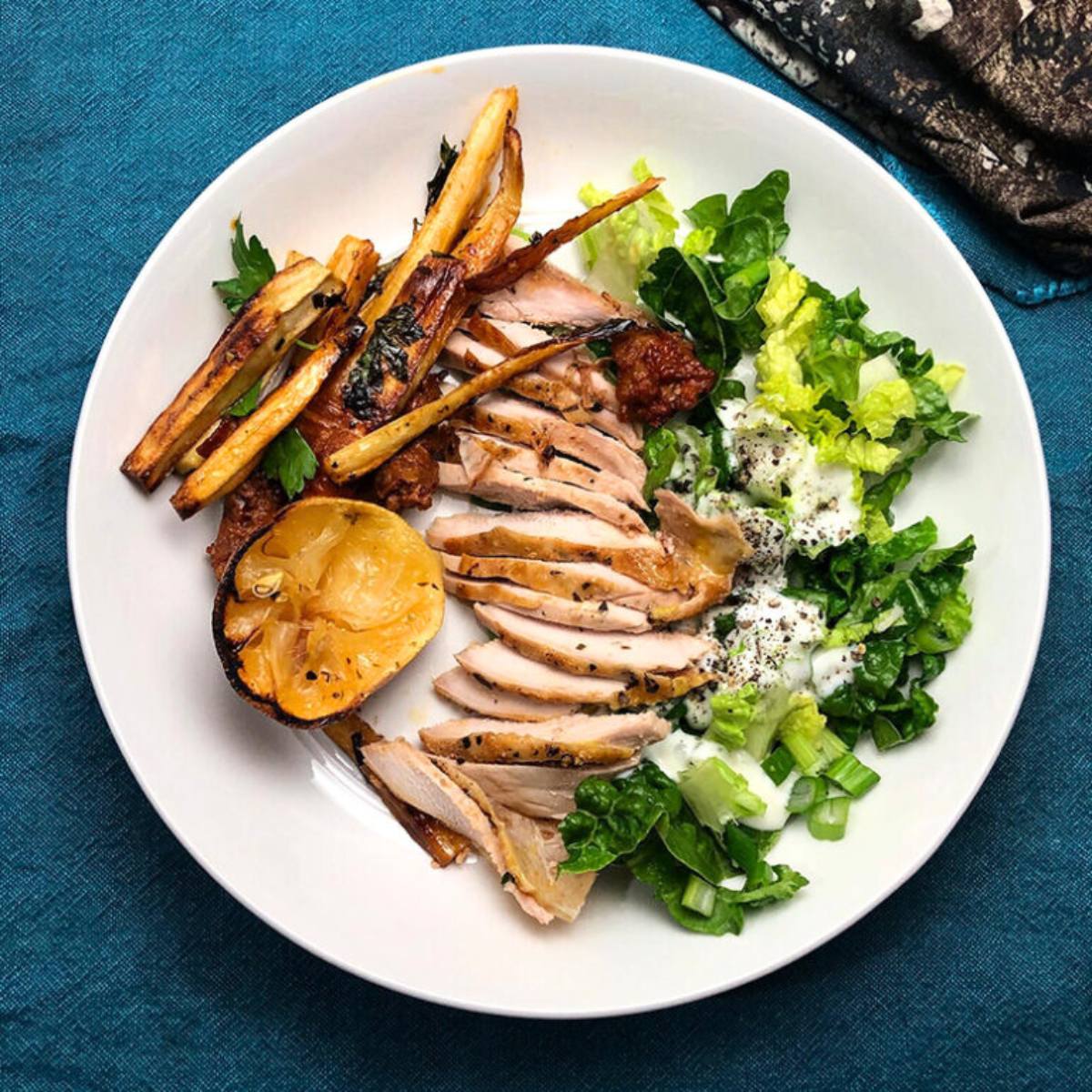 A white plate with a healthy and colorful meal. The plate features sliced roasted pheasant, roasted parsnips, a green salad with a creamy dressing, and a lemon wedge.