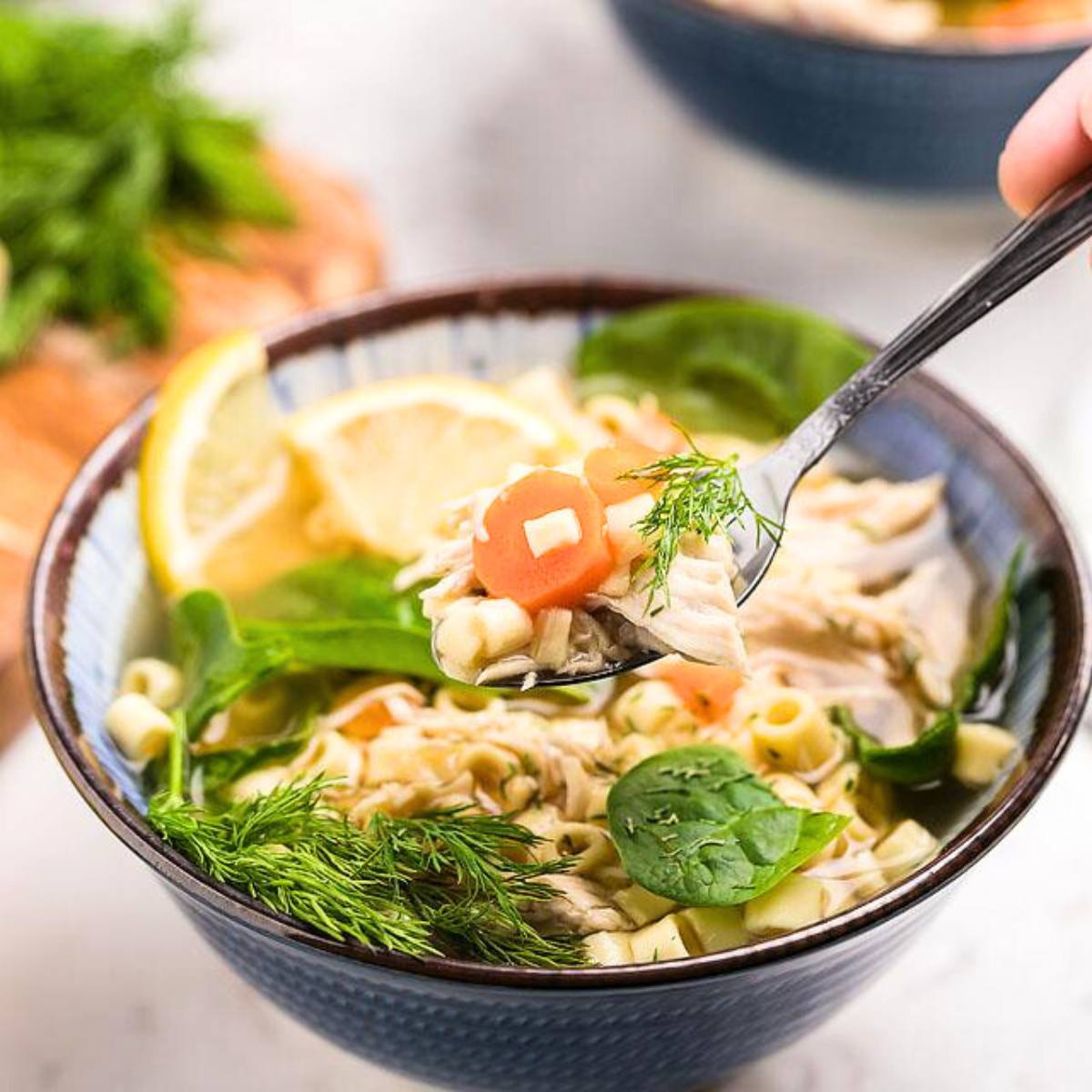 A bowl of classic chicken noodle soup with carrots, spinach, and a lemon wedge.