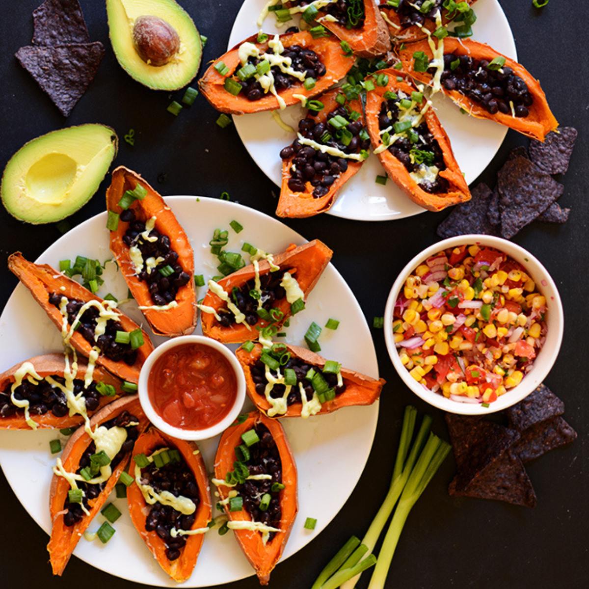 A top-down view of two plates of loaded sweet potato boats filled with black beans, salsa, avocado, and green onions. There's also a bowl of corn salsa and tortilla chips on the side.