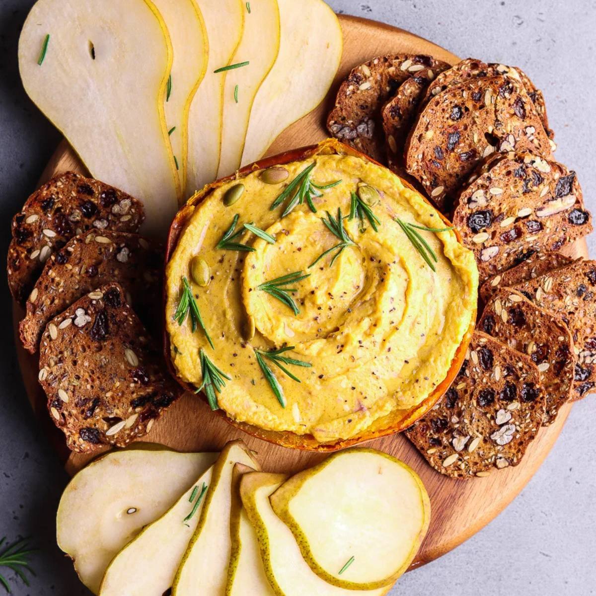 A halved pumpkin filled with a creamy dip, surrounded by sliced pears and crackers on a wooden board.