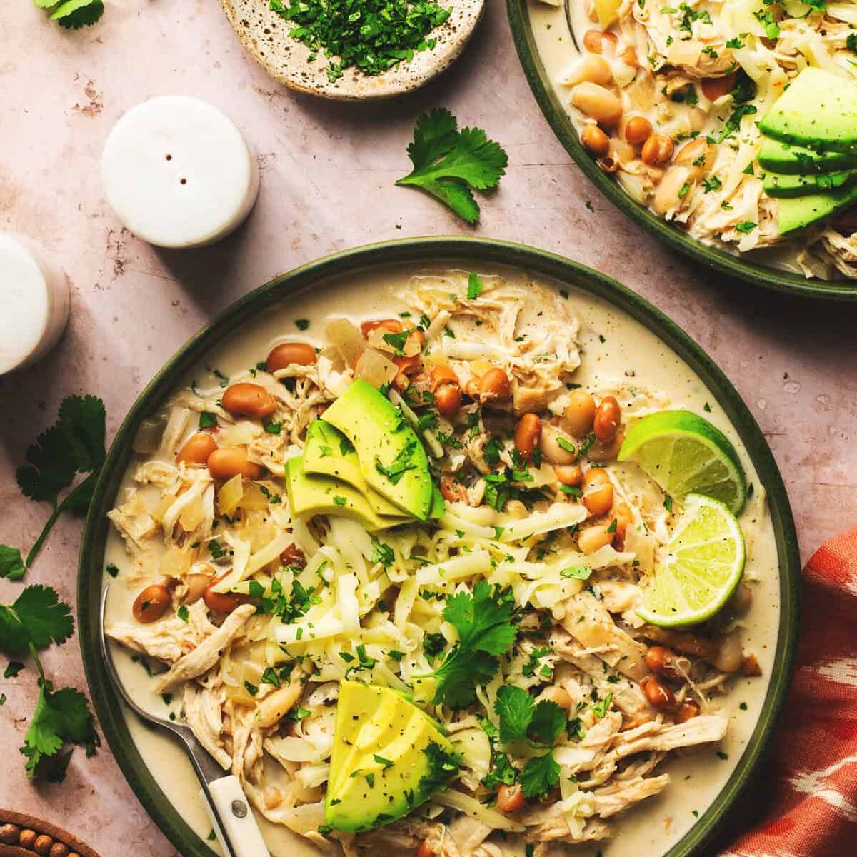 Two bowls of creamy white chicken chili with beans, avocado, cheese, cilantro, and lime wedges.