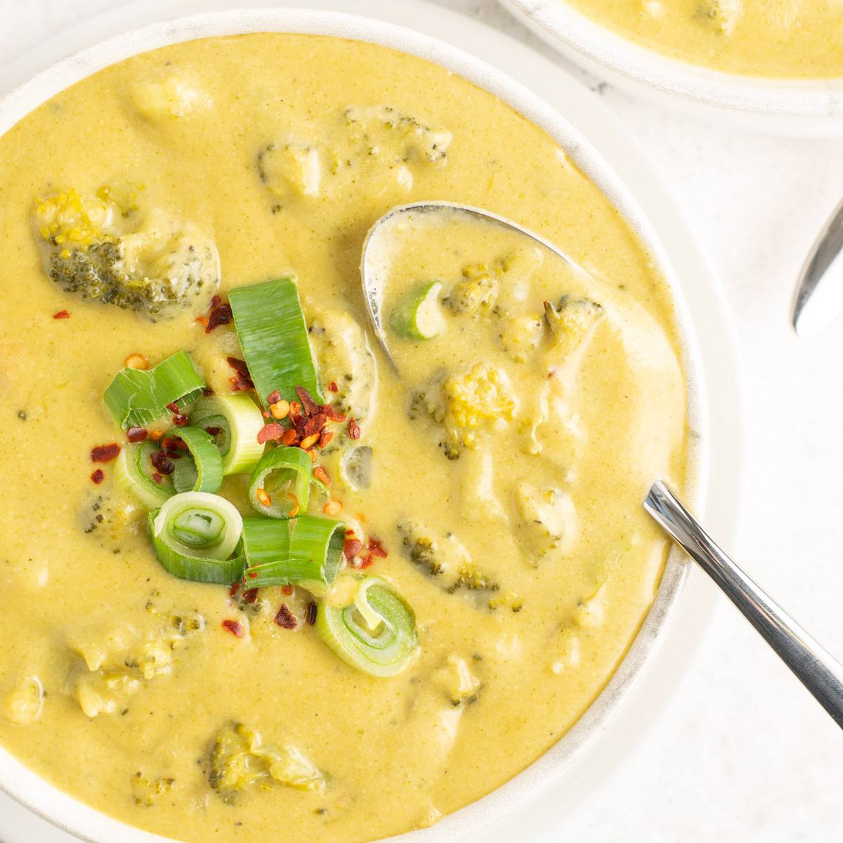 A white bowl filled with a creamy, golden broccoli cheddar soup. The soup is topped with green onions and red pepper flakes. A spoon is scooping up a bite of the soup.