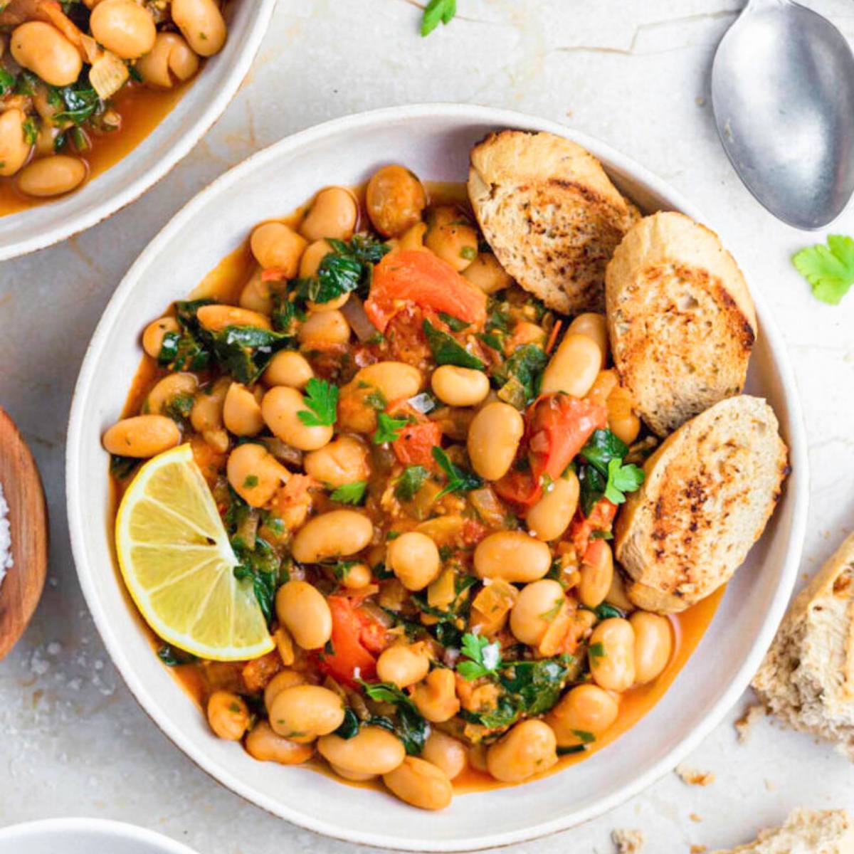 A white bowl filled with a hearty white bean stew. The stew is made with white beans, tomatoes, spinach, and herbs, and is garnished with a lemon wedge and toasted bread.