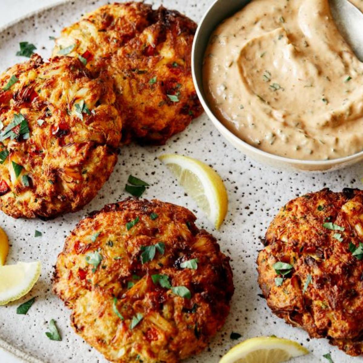 A plate of golden-brown crab cakes with a side of creamy dipping sauce and lemon wedges.