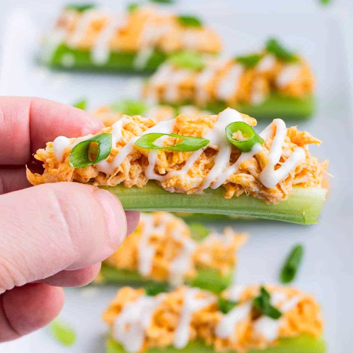 A hand holding a celery stick filled with creamy buffalo chicken dip, topped with green onions.