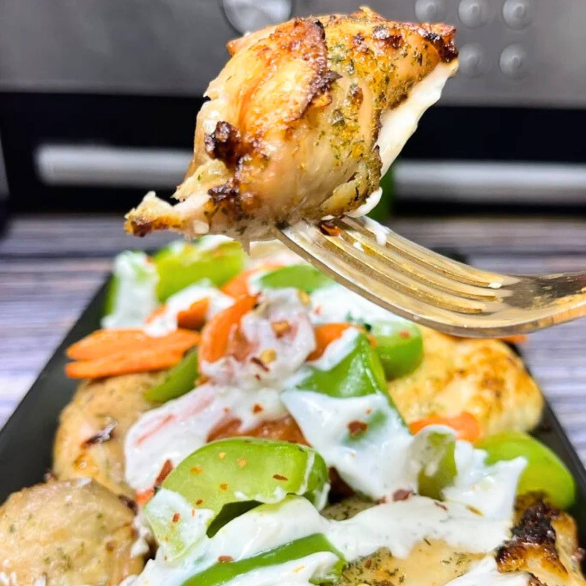 A fork holding a piece of roasted chicken with a side of roasted vegetables and a creamy sauce.