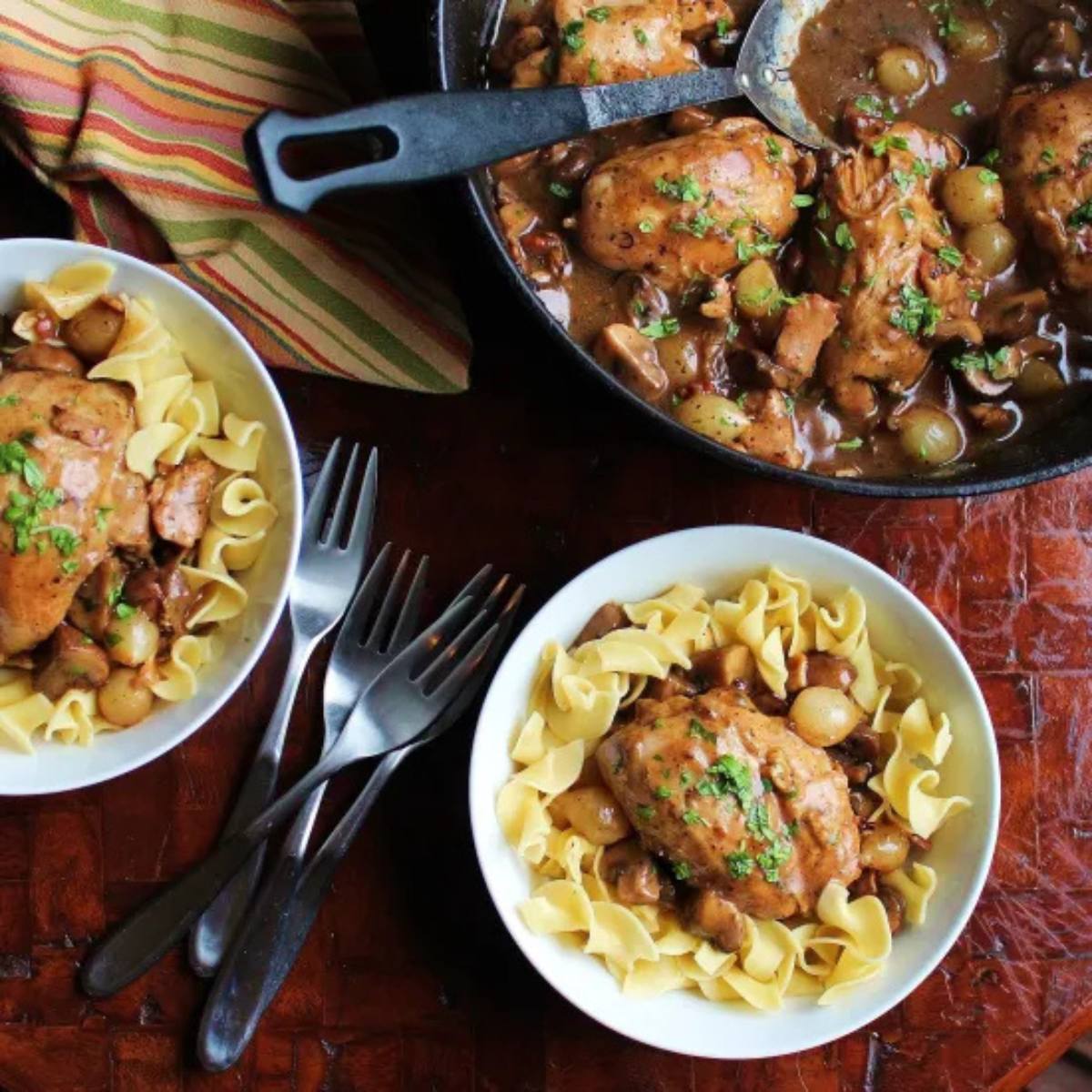 A cast iron skillet filled with a rich, brown sauce with chicken, grapes, and mushrooms. Two white bowls filled with the same dish and egg noodles are also visible.