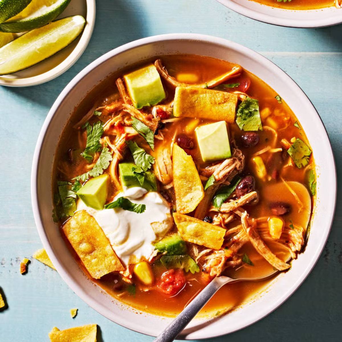 The soup itself is a vibrant orange color and filled with chicken, beans, corn, and tomatoes. The toppings include avocado slices, sour cream, tortilla chips, and cilantro.