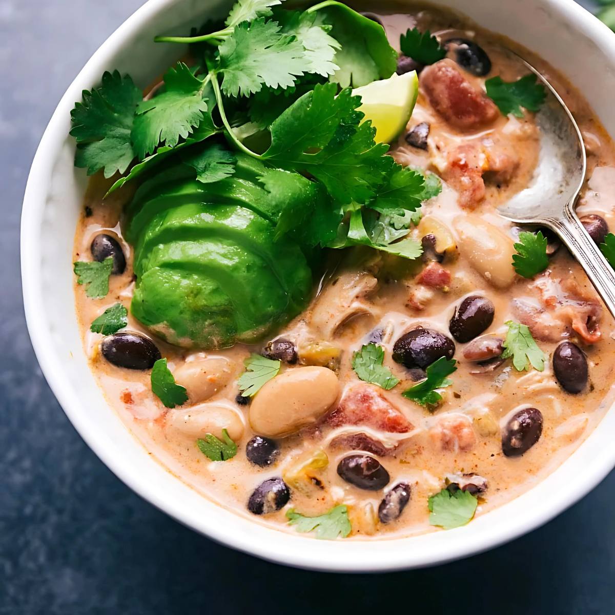 A bowl of creamy white chicken chili with beans, avocado, cilantro, and a lime wedge.