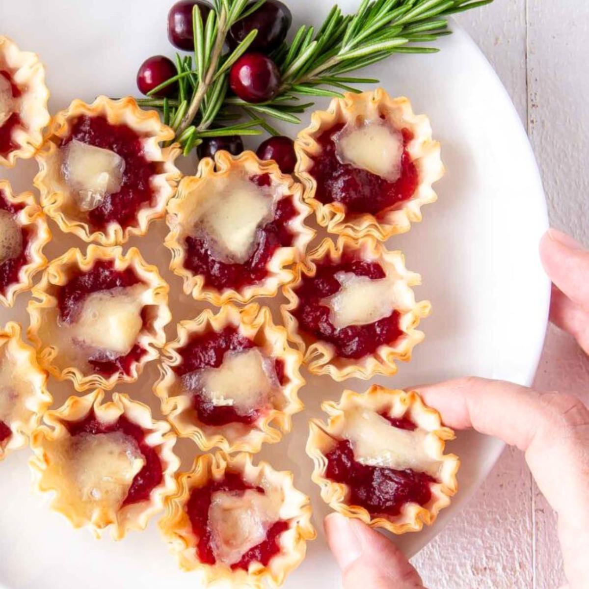 A plate of small, golden-brown pastry cups filled with cranberry sauce and topped with melted brie cheese.