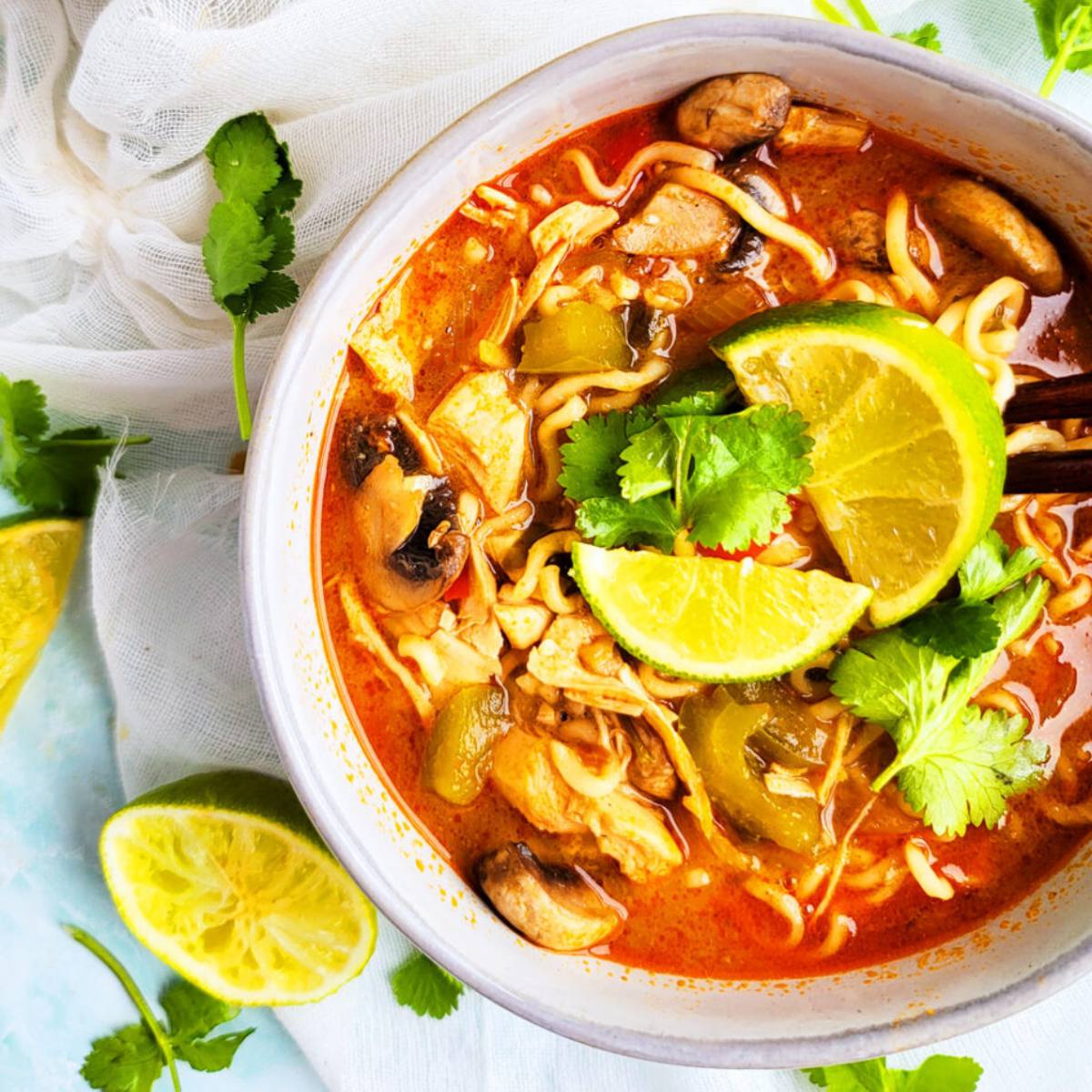 A bowl of spicy Thai noodle soup with chicken, mushrooms, and vegetables. The soup is a vibrant red color and is topped with lime wedges, cilantro, and noodles.