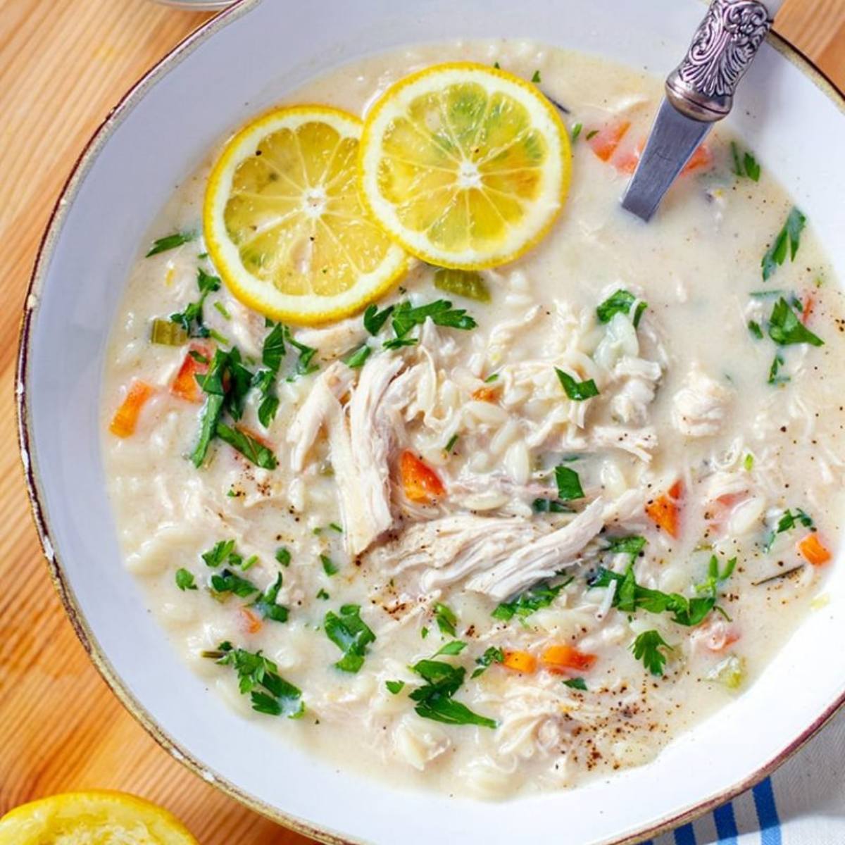 A white bowl filled with a creamy lemon chicken soup. The soup is made with shredded chicken, rice, vegetables, and is garnished with lemon slices and fresh parsley.