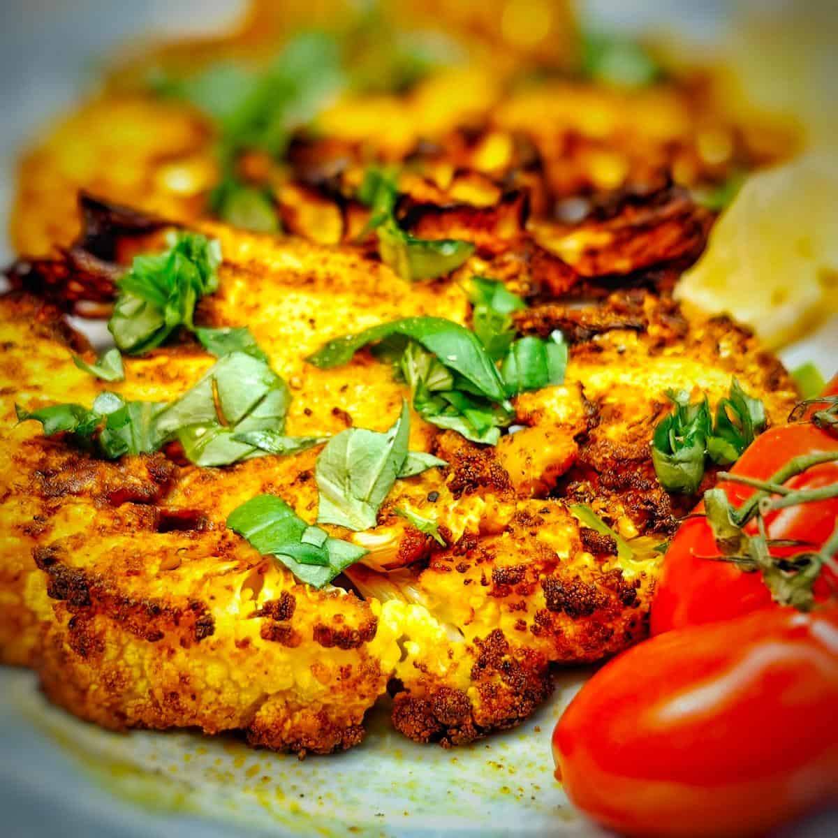 A close-up of a plate of golden-brown roasted cauliflower steaks topped with fresh basil leaves, served alongside roasted cherry tomatoes.