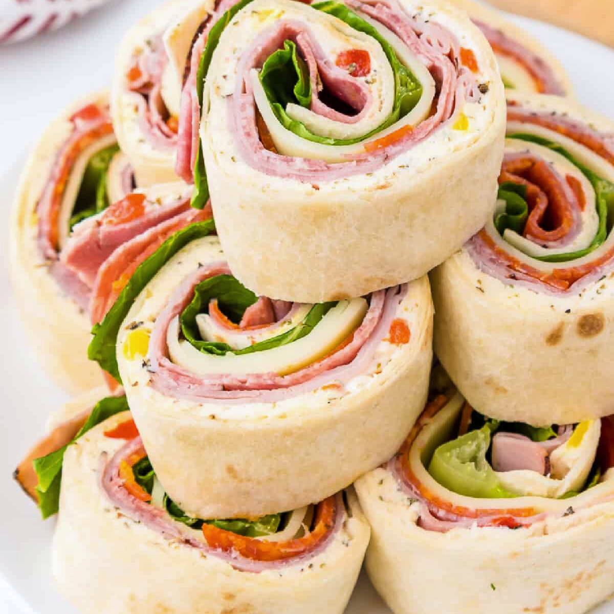 A plate of rolled-up tortilla wraps filled with ham, salami, cheese, lettuce, and peppers.