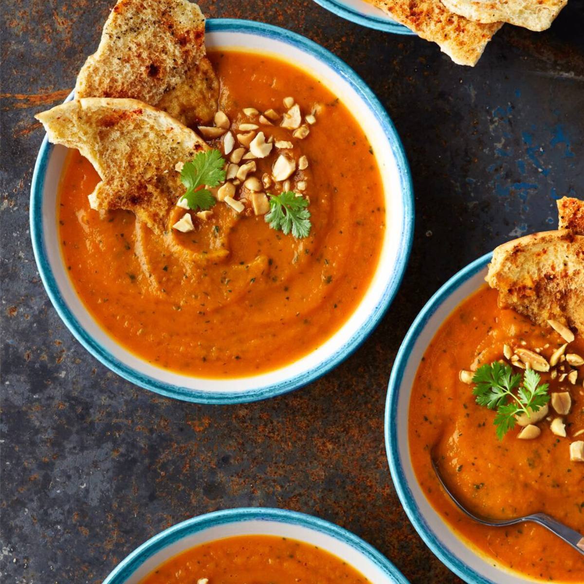 Three bowls of vibrant orange soup topped with toasted pita bread, peanuts, and cilantro. 