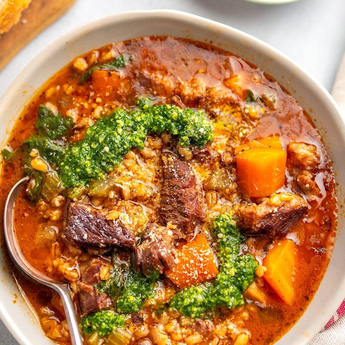 A white bowl filled with a steaming bowl of barley soup. The soup is made with beef, carrots, and other vegetables, and is topped with a dollop of green pesto. A spoon rests in the bowl.