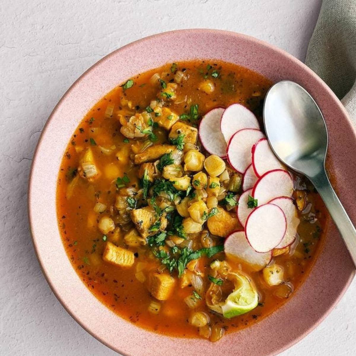 A bowl of pozole, a traditional Mexican soup with hominy, pork, and a variety of toppings. The soup is a vibrant red color and is topped with radishes, cilantro, and lime wedges.