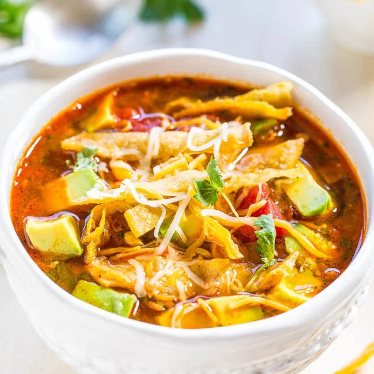 A white bowl filled with a hearty chicken tortilla soup. The soup is topped with shredded cheese, tortilla strips, avocado, and cilantro.