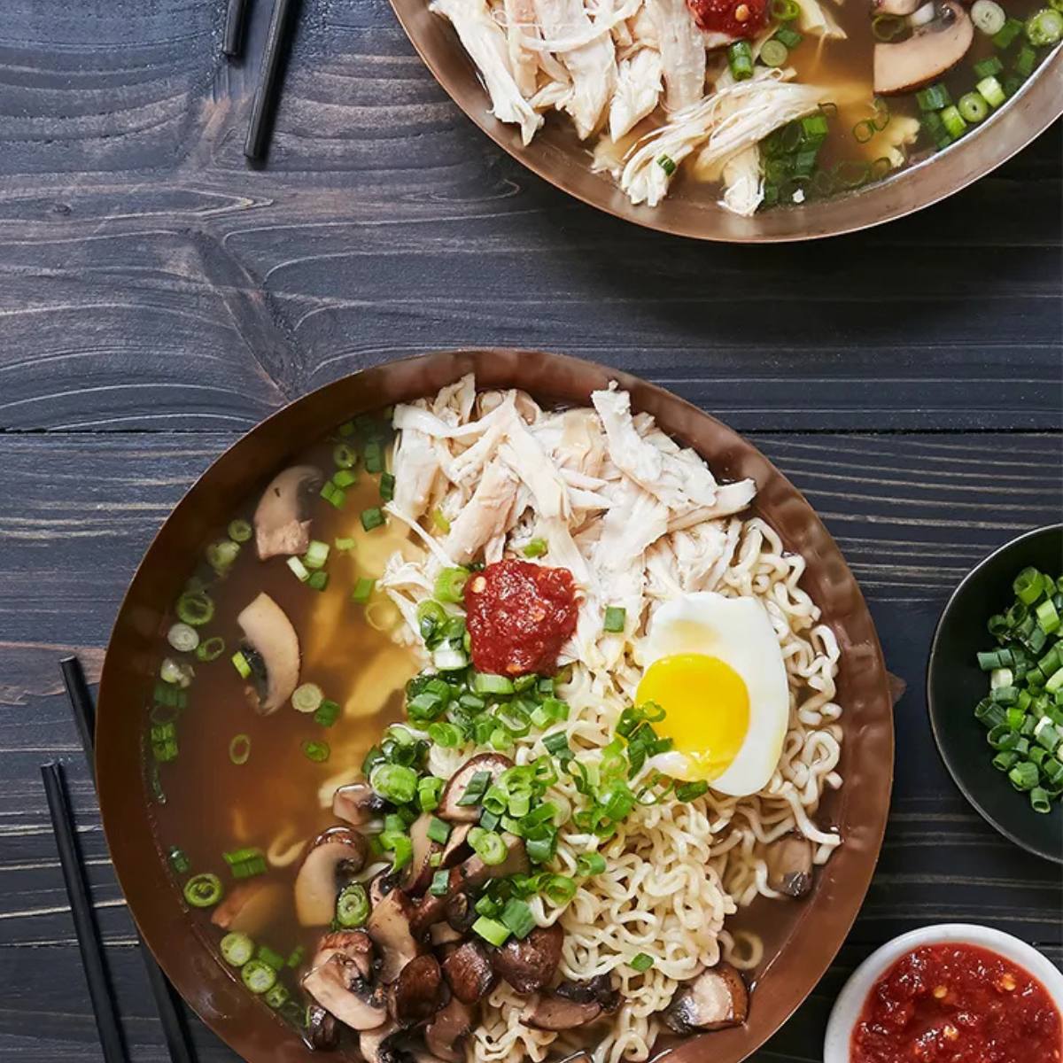 Two bowls of ramen noodles topped with shredded chicken, sliced mushrooms, green onions, a soft-boiled egg, and a drizzle of chili oil. Chopsticks and a small bowl of chili oil are also visible.