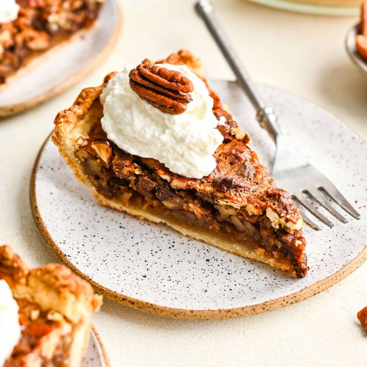 A slice of pecan pie on a speckled plate with a dollop of whipped cream and a pecan on top.