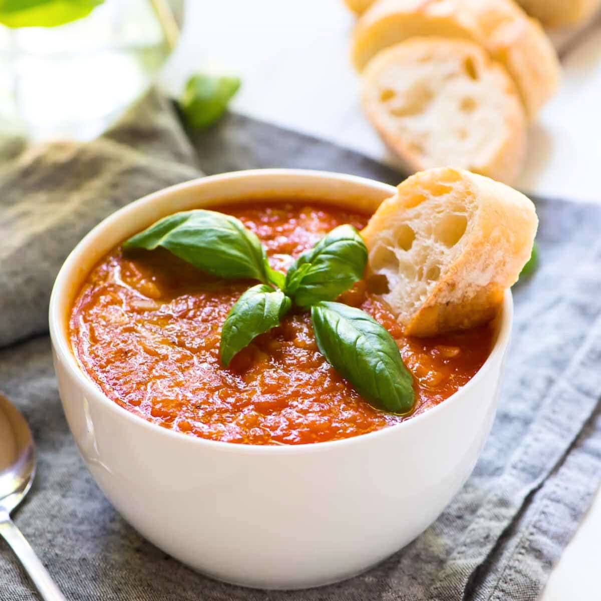 A bowl of creamy tomato soup with fresh basil and a piece of crusty bread. The soup is a vibrant red color and has a smooth texture.