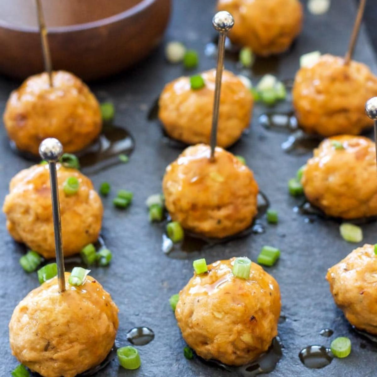 A close-up of several small, golden-brown meatballs on skewers, drizzled with a sweet and savory glaze and garnished with green onions.