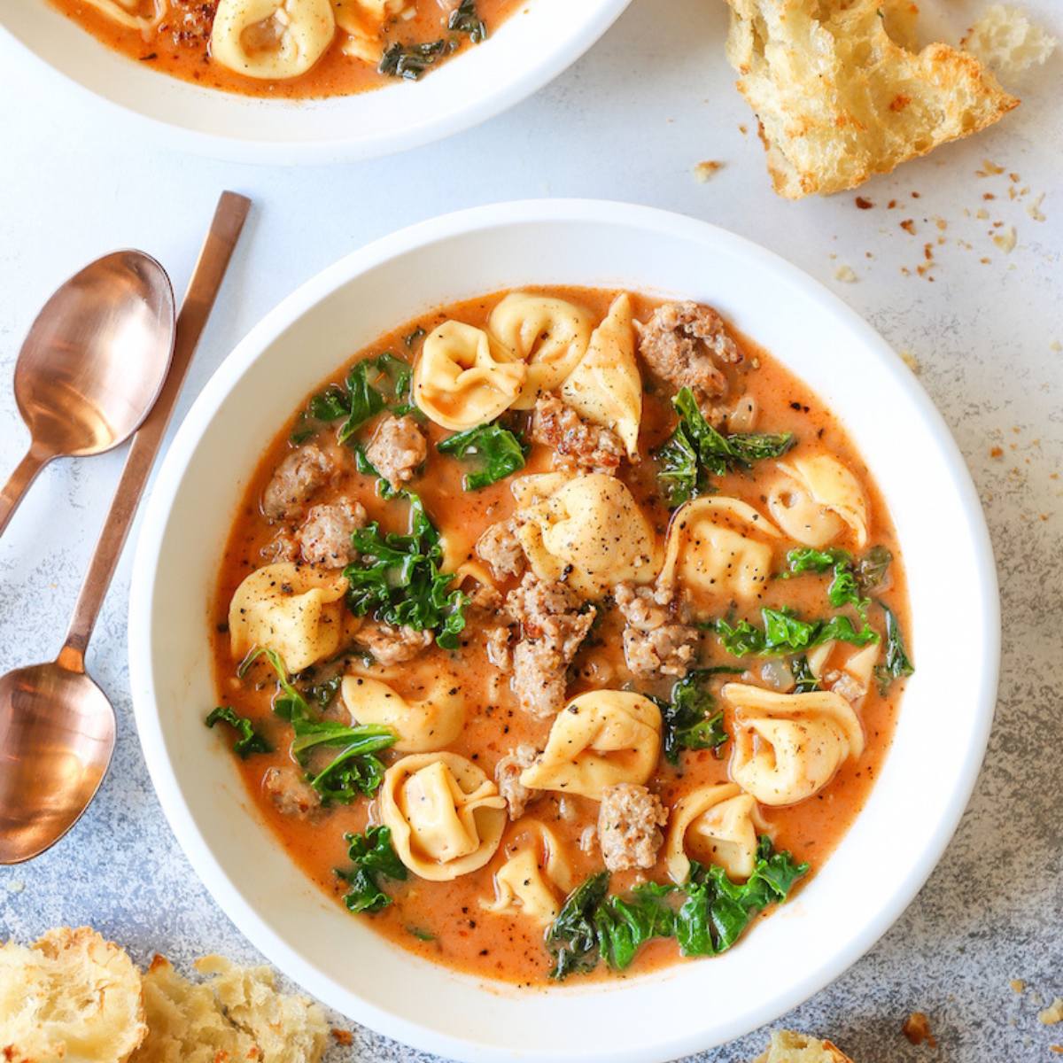 A white bowl filled with a hearty tortellini soup. The soup is made with tortellini, sausage, kale, and a tomato-based broth. A piece of crusty bread is visible in the background.