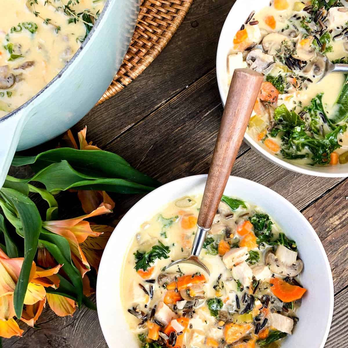 Two white bowls filled with a creamy wild rice soup. The soup is made with wild rice, carrots, mushrooms, and kale, and is topped with fresh herbs.