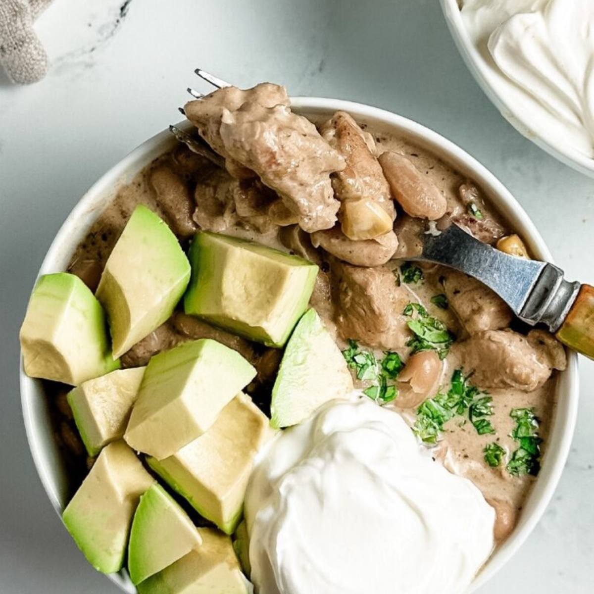 A white bowl filled with a creamy white chicken chili. The chili is topped with avocado slices, dollops of sour cream, and fresh cilantro. A fork is taking a bite of the chili.