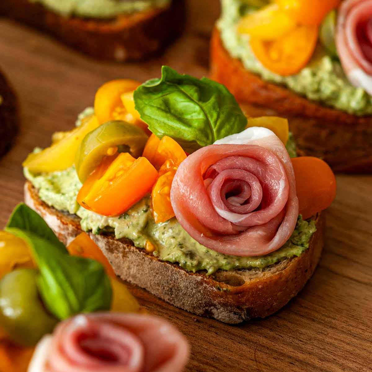 A close-up of an open-faced sandwich on toasted bread with a green spread, topped with cherry tomatoes, olives, a rose-shaped prosciutto slice, and fresh basil leaves.