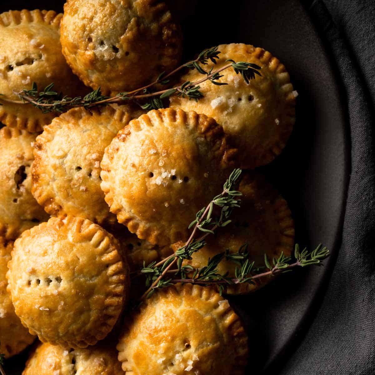 A close-up of several golden-brown mini meat pies on a dark plate, garnished with fresh thyme.