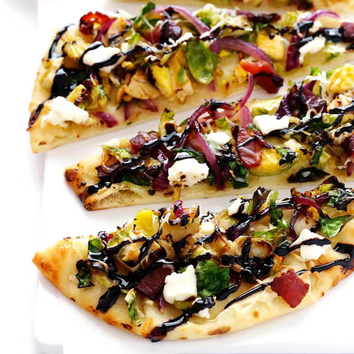 Three slices of flatbread topped with roasted Brussels sprouts, bacon, onions, and goat cheese, drizzled with balsamic glaze.