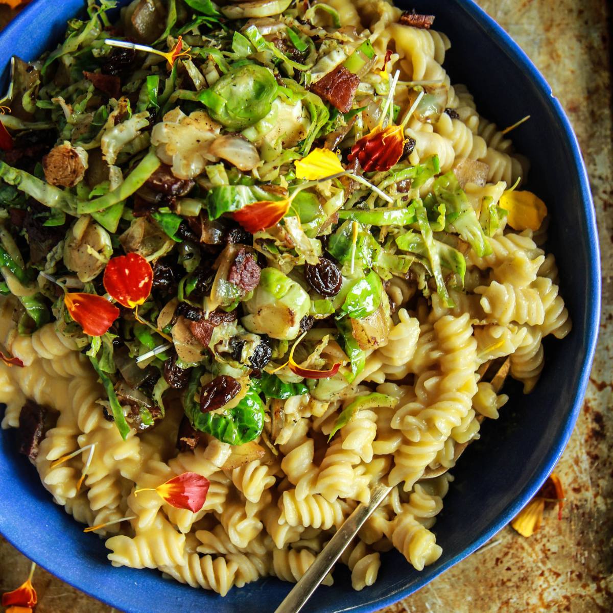 A bowl of pasta with a creamy sauce, topped with sautéed Brussels sprouts, bacon, and edible flowers.