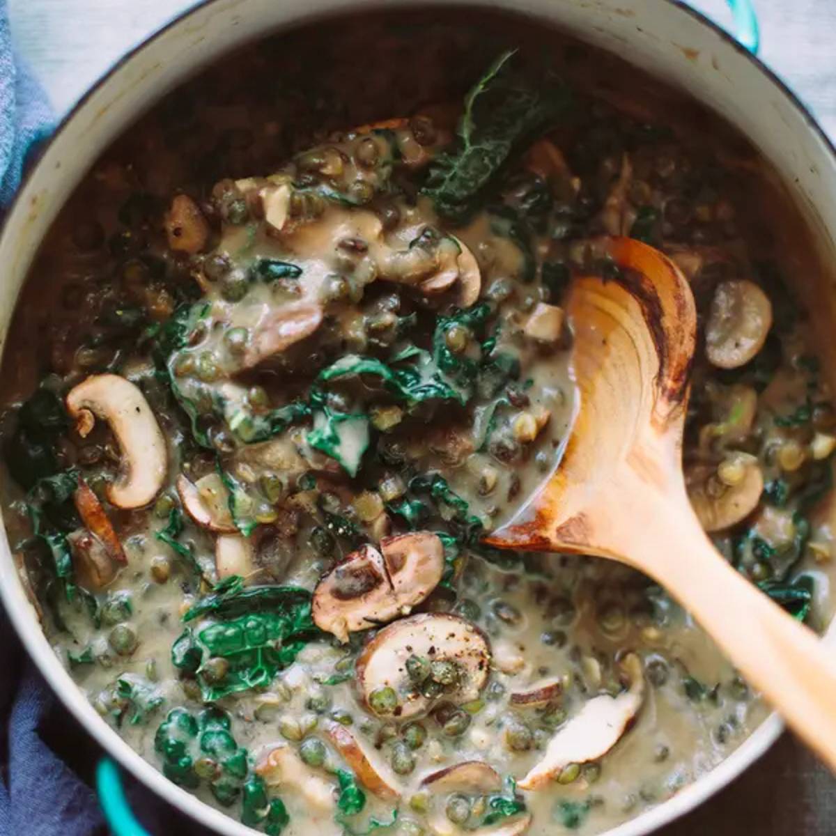 A pot of creamy lentil soup with mushrooms, kale, and a wooden spoon.