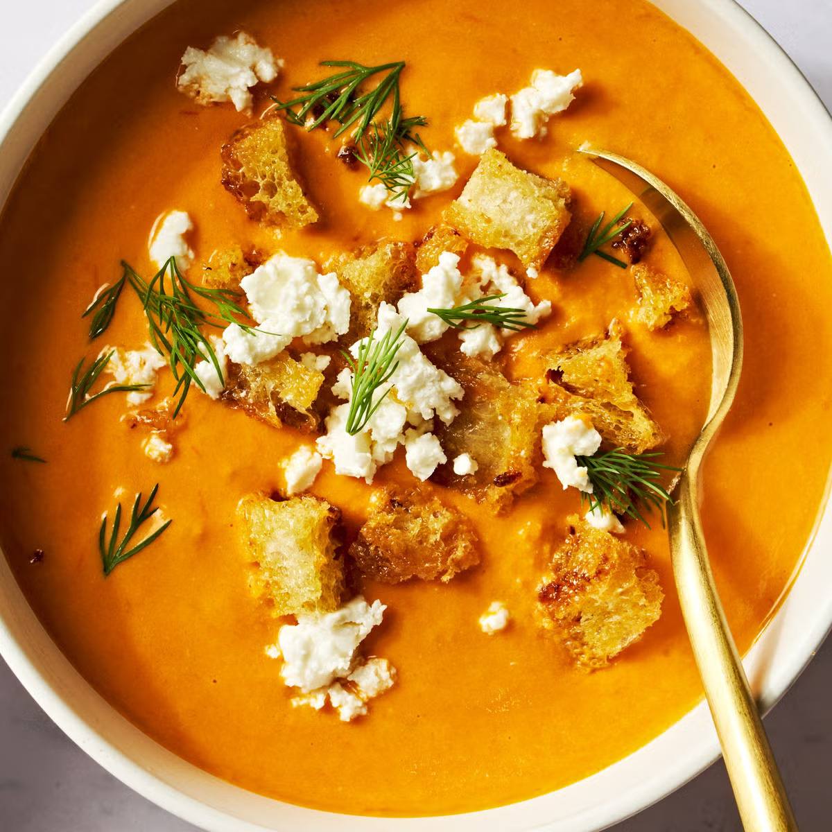 A white bowl filled with a vibrant red pepper soup. The soup is topped with crumbled feta cheese, croutons, and fresh dill. A golden spoon rests in the bowl.

