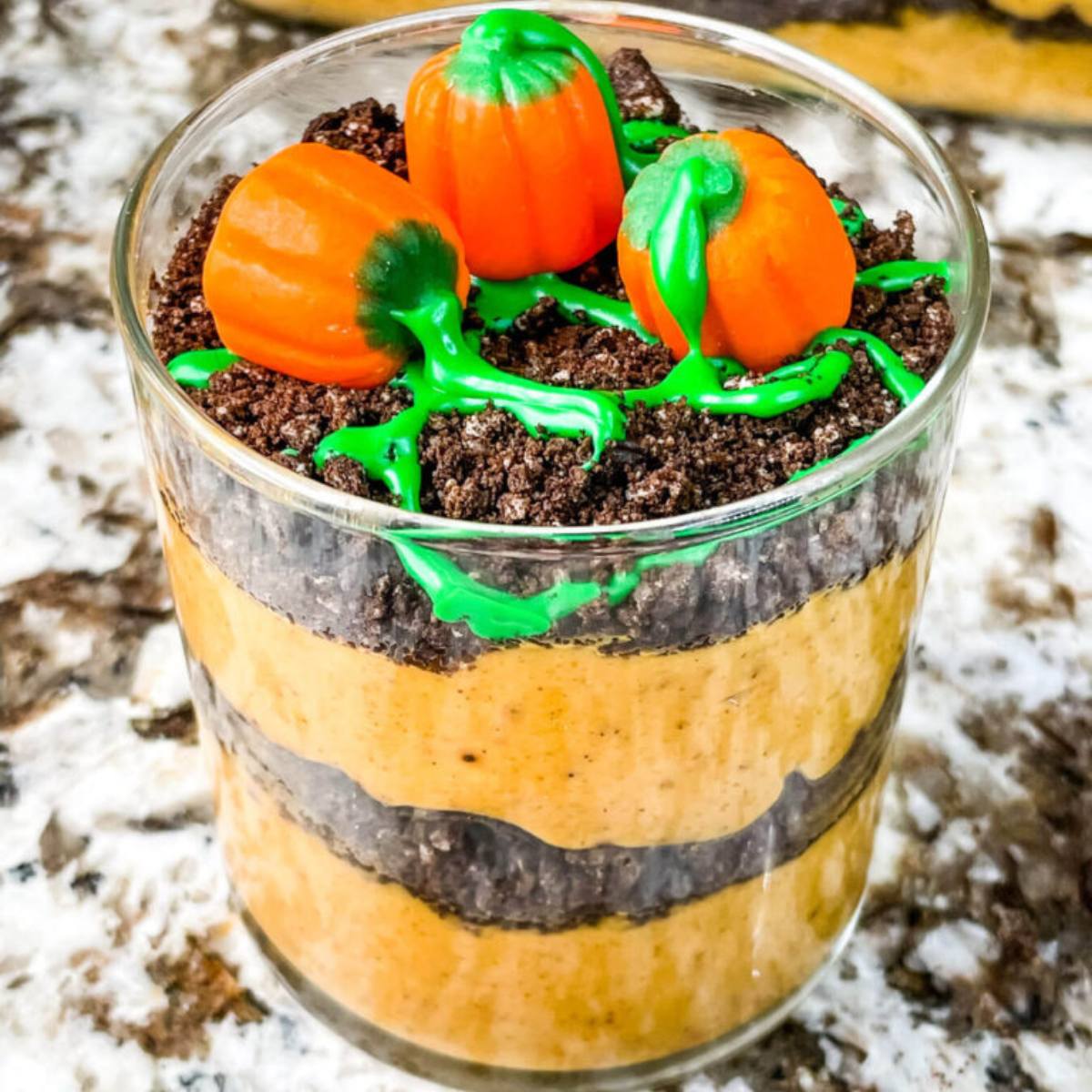 A glass jar filled with layers of pumpkin pudding and crushed Oreo cookies. The top layer is decorated with green frosting and orange pumpkin candies.