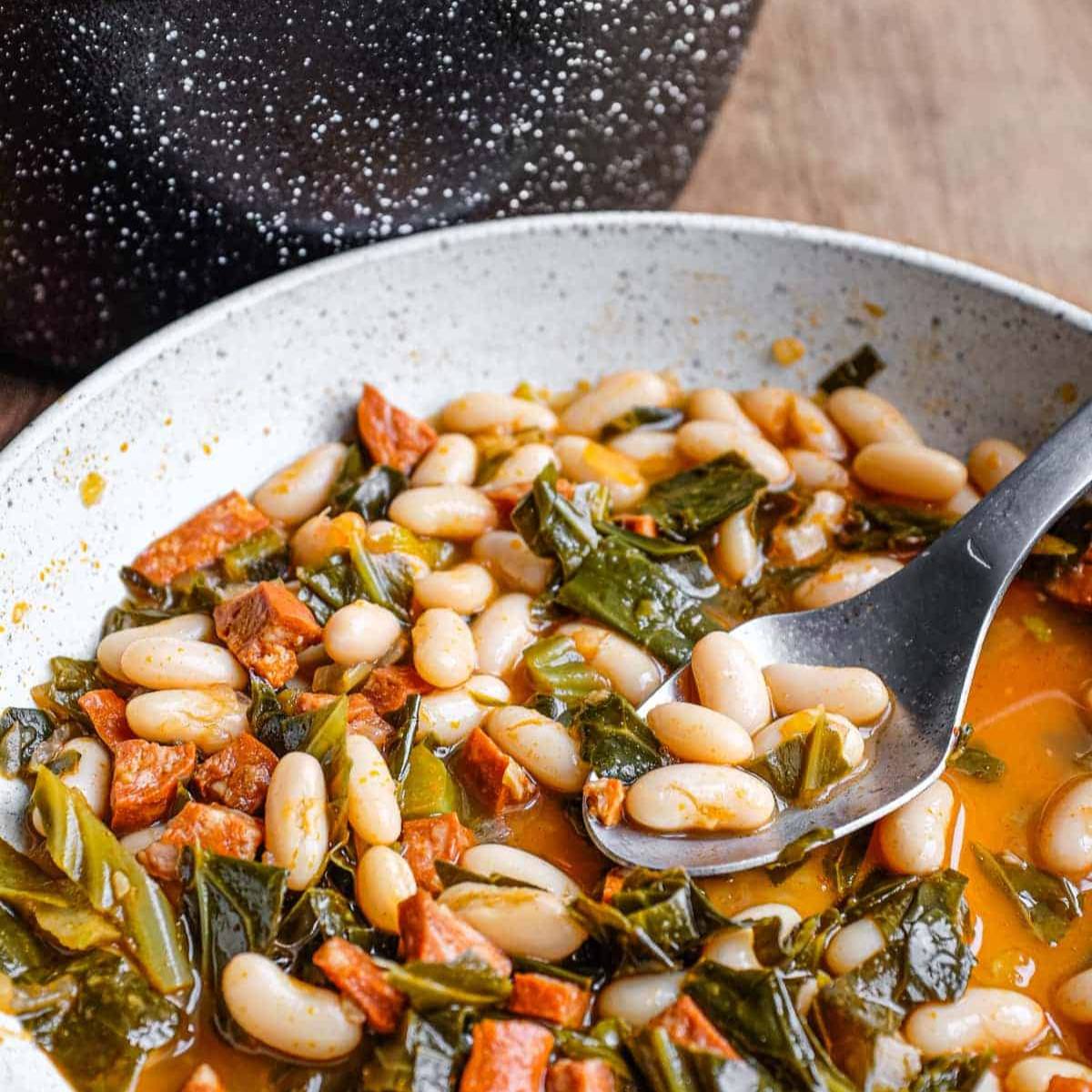 A white bowl filled with a hearty soup of white beans, greens, and sausage. The soup is simmered in a flavorful broth and is ready to be served.