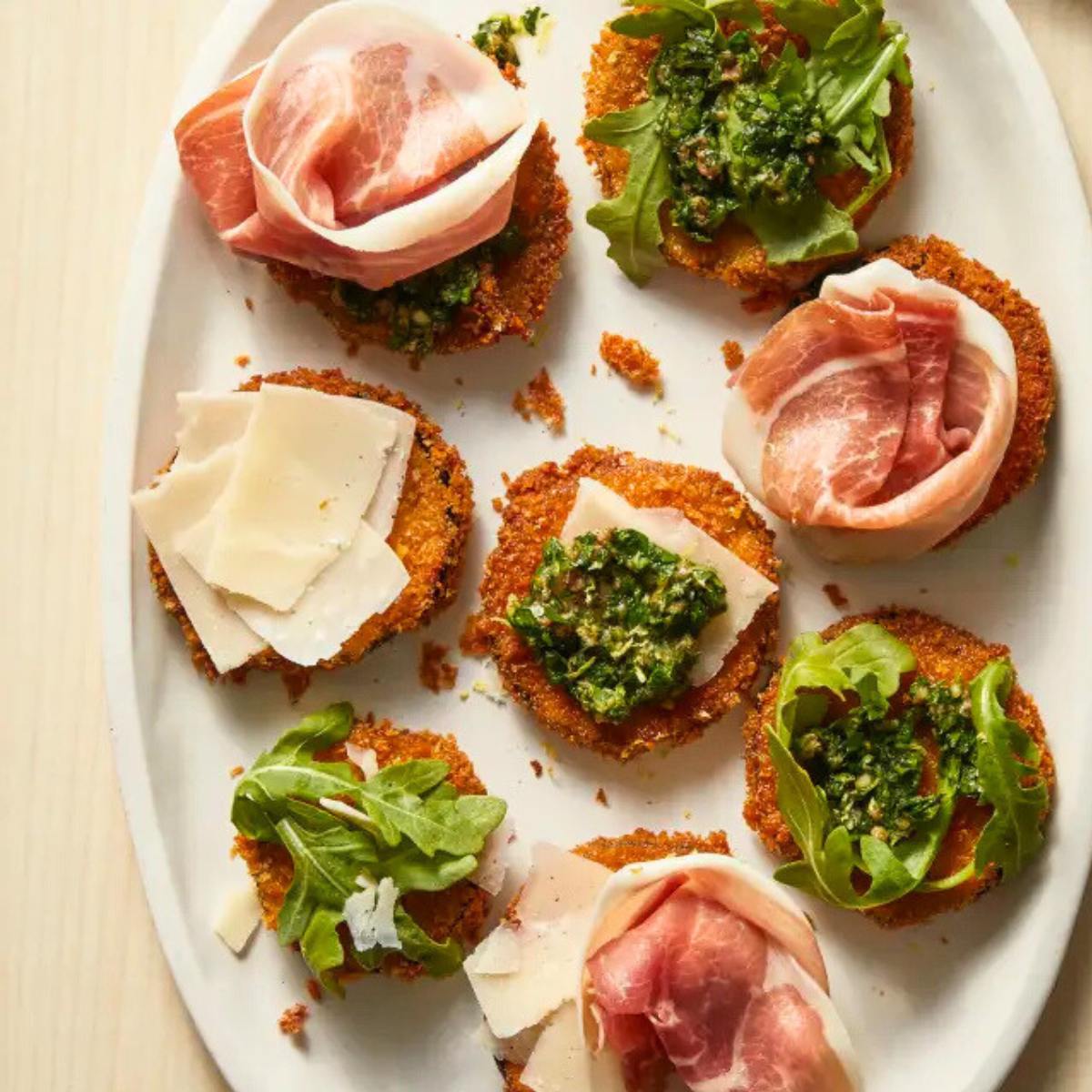 A plate of crispy fried eggplant rounds topped with various toppings, including prosciutto, arugula, Parmesan cheese, and pesto.