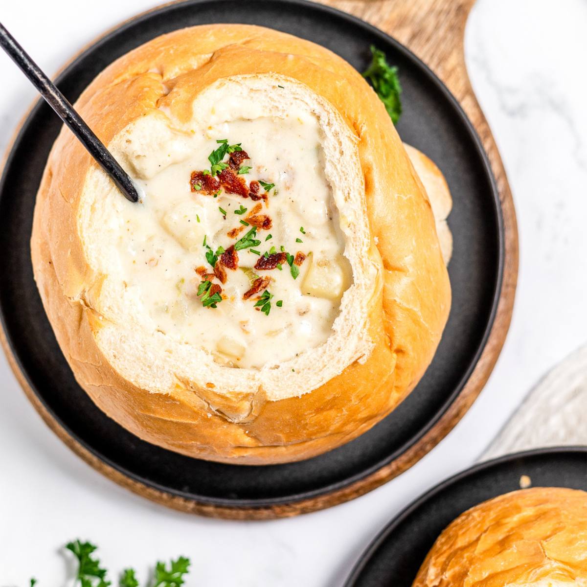 A top-down view of a bread bowl filled with creamy clam chowder. The chowder is topped with crispy bacon bits, fresh parsley, and a spoon.