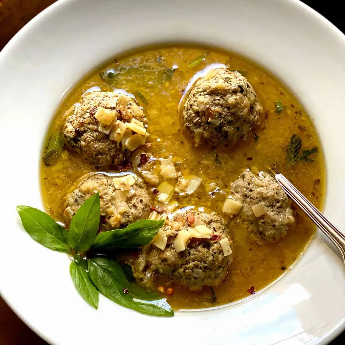 A white bowl filled with a golden broth. Several meatballs are floating in the broth, and the dish is garnished with fresh basil leaves.