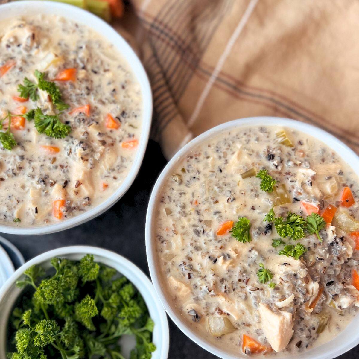 Two white bowls filled with a creamy chicken and wild rice soup. The soup is topped with fresh parsley and is served with a small bowl of additional parsley on the side.